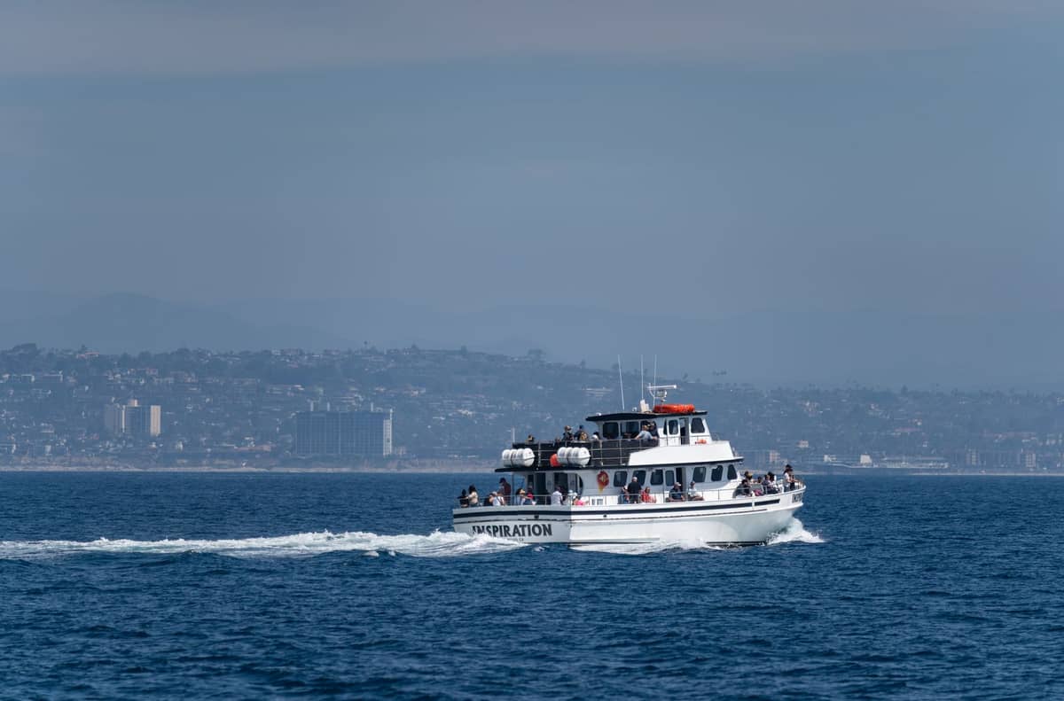 Ein mittelgroßes Touristenboot mit Passagieren fährt vor der Küste, mit der Küstenlinie und der Stadt San Diego im Hintergrund unter einem bewölkten Himmel.