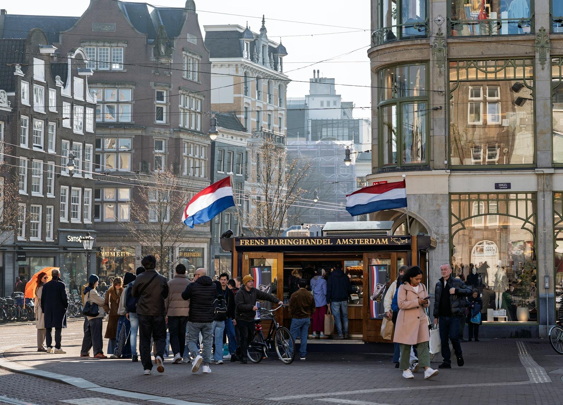 Animada escena callejera en Ámsterdam con personas reunidas en un puesto tradicional de arenque, banderas holandesas y edificios históricos al fondo.