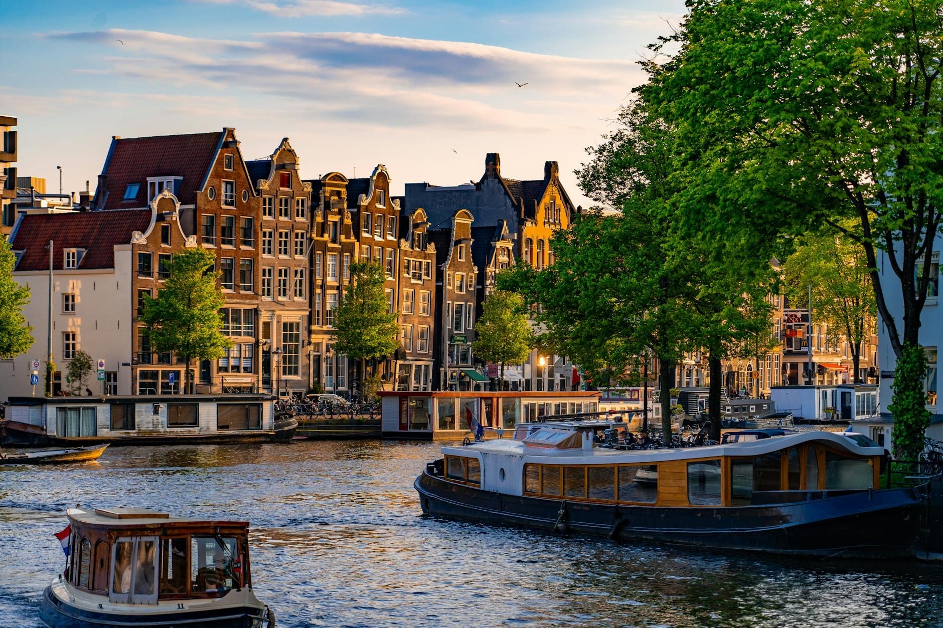 Vista clásica del canal de Ámsterdam con casas históricas de hastiales, barcos-casa y botes bajo cielos soleados, capturando perfectamente el famoso Canal Ring de la ciudad.