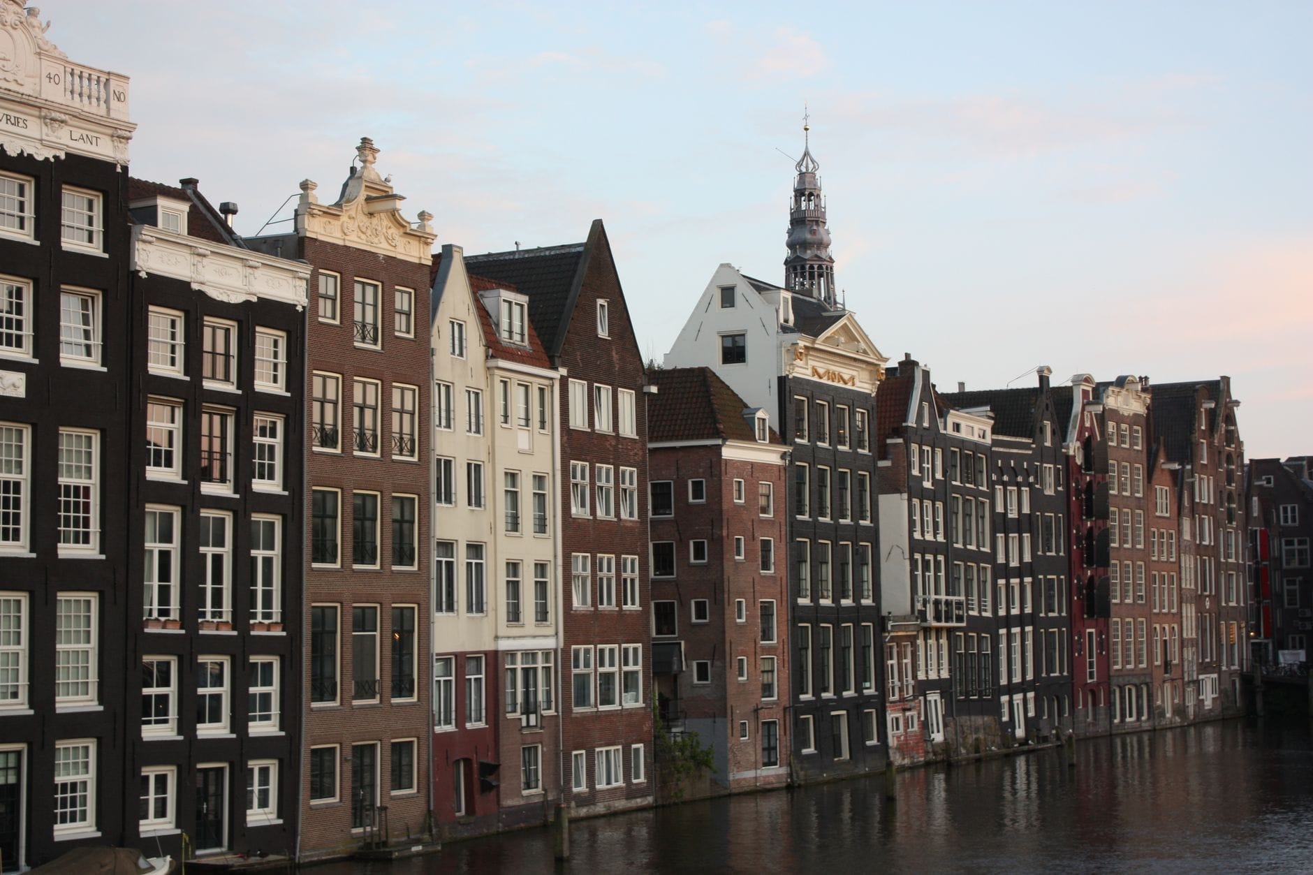 Casas típicas de canal de Ámsterdam con elaborados frontones y fachadas estrechas bordean un frente de agua, con la aguja de una iglesia visible al fondo bajo un cielo azul.