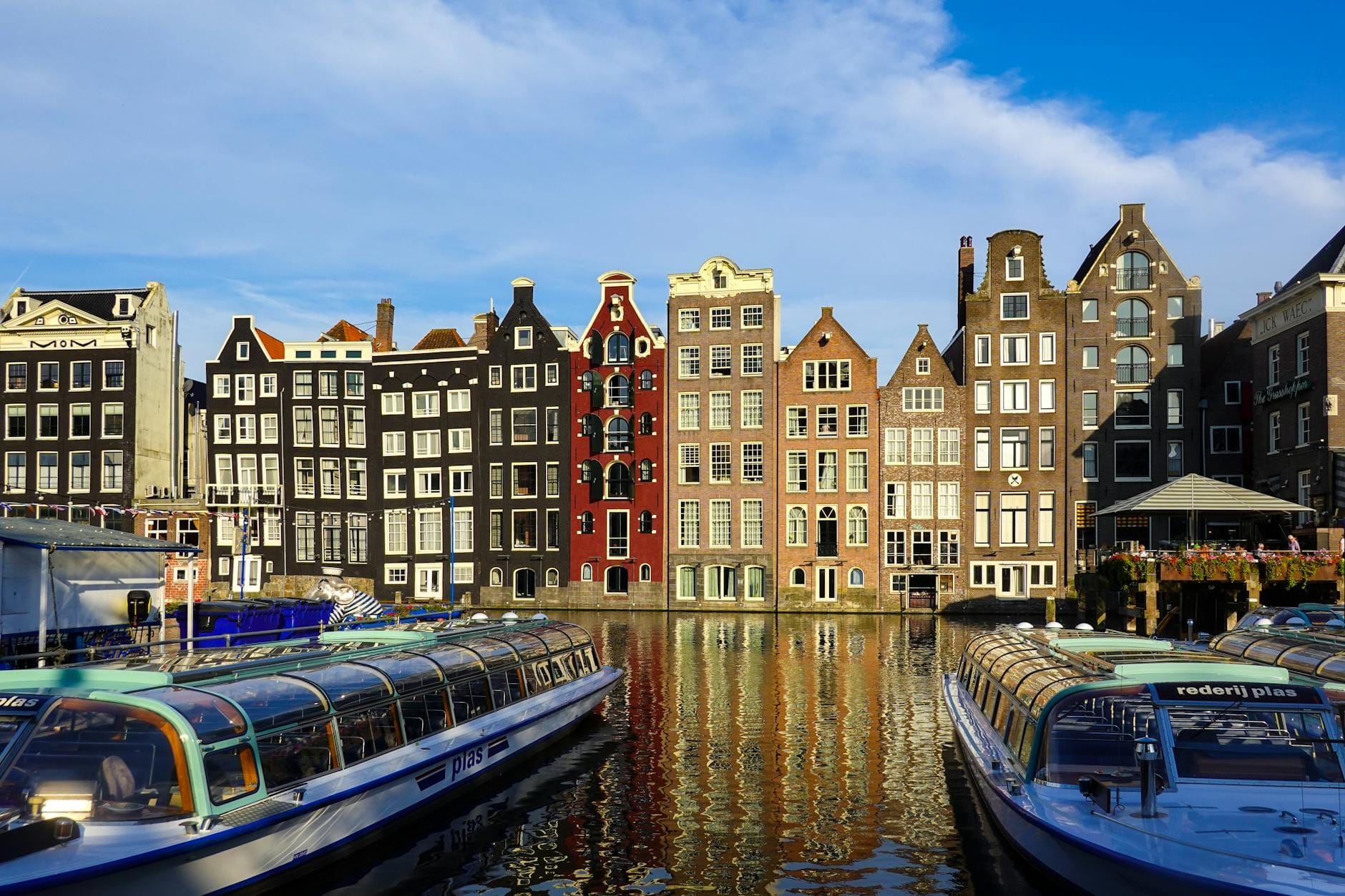 Escena clásica de los canales de Ámsterdam con estrechas casas de colores y frontones reflejadas en el agua, flanqueadas por barcos turísticos con techo de cristal bajo un cielo azul intenso.