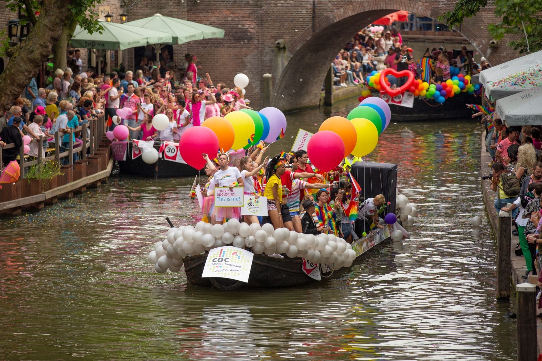 Barca de canal decorada con globos de arcoíris y personas celebrando durante un desfile del orgullo en Ámsterdam, con multitudes observando desde ambos lados del canal.