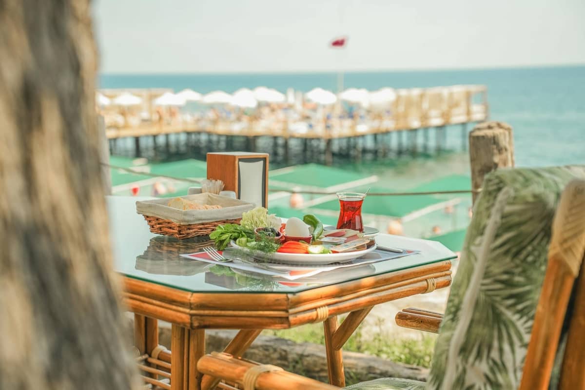 Breakfast table with Turkish tea, vegetables, and bread set outdoors overlooking the sea in Antalya with a pier and umbrellas in view.