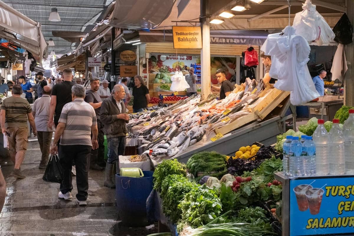 Bustling market scene in Antalya featuring locals, fresh fish, vegetables, and Turkish market signage under covered stalls.