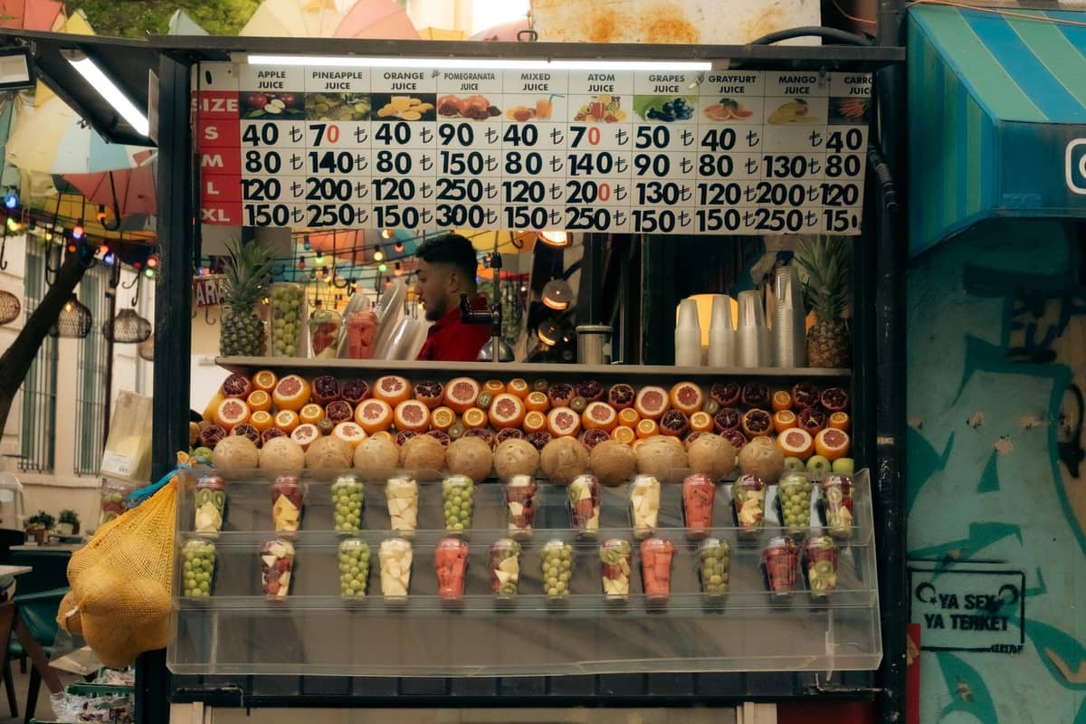 A colorful juice stand in a street setting with fresh oranges, pomegranates, pineapples, and cups of mixed fruit, offering a variety of drinks in Antalya.