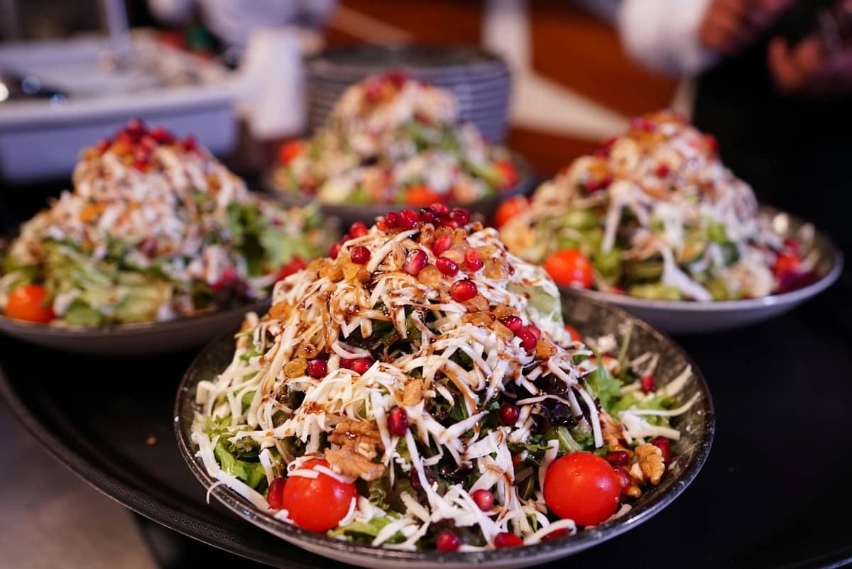 Four plates piled high with Turkish salads, including pomegranate seeds, walnuts, cherry tomatoes, and shredded white cheese. Vibrant and family-style presentation in a restaurant setting.