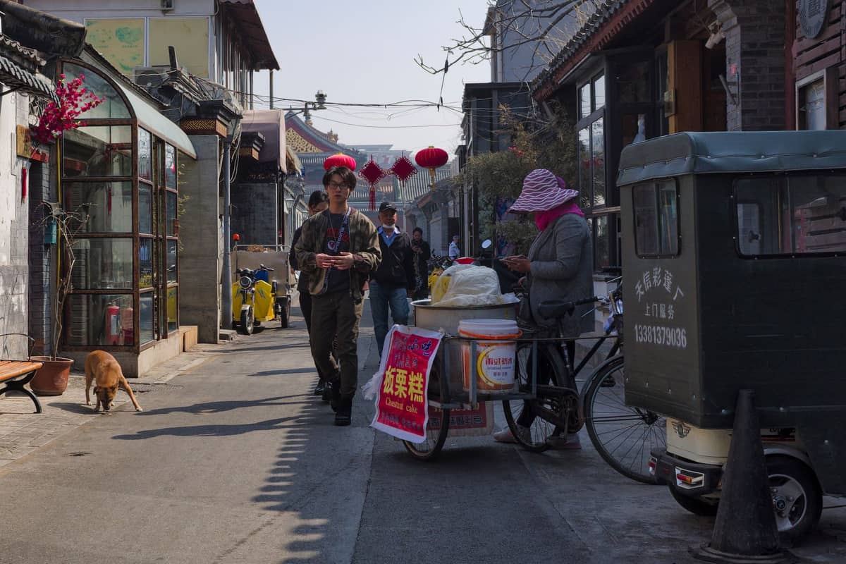 Straßenhändler mit einem Essenskarren, der Frühstück in einem traditionellen Hutong in Beijing verkauft, umgeben von Einheimischen und historischen Gebäuden der Gasse.