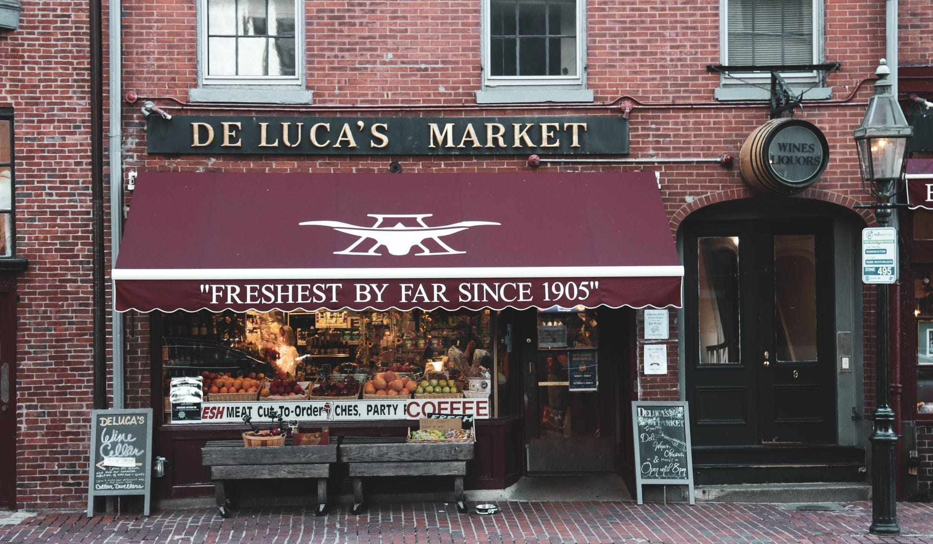 Fachada histórica do De Luca's Market em Boston, com toldo vermelho, frutas frescas na vitrine e aquela energia acolhedora de bairro numa rua de paralelepípedos.