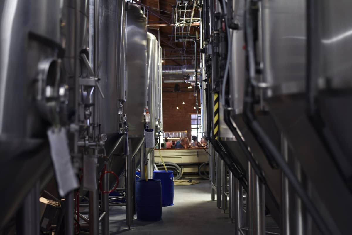 Große Stahl-Brautanks säumen einen Korridor, im Hintergrund sitzen Menschen an Tischen, was auf eine aktive Brauerei hindeutet.