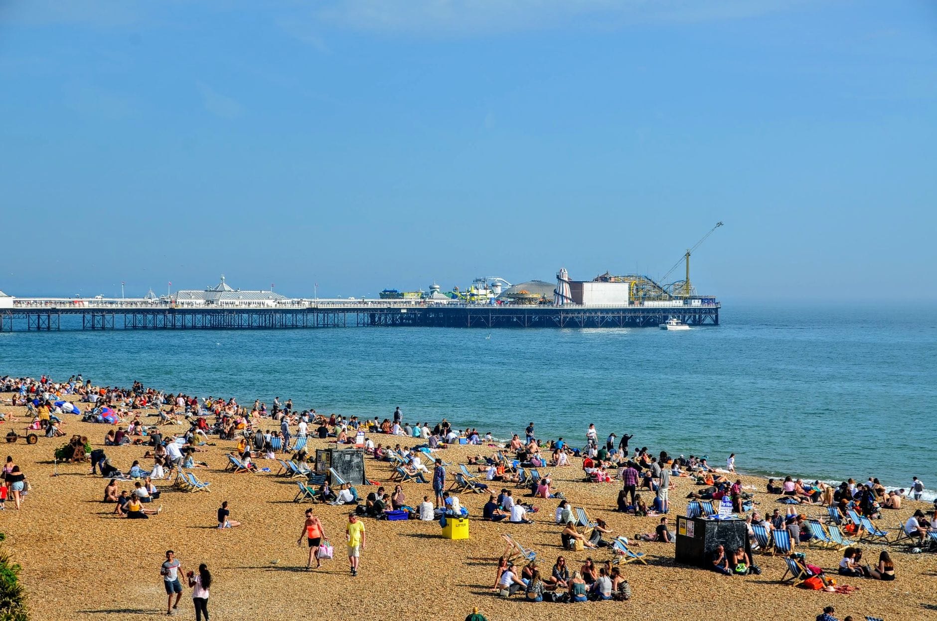 Animata scena estiva sulla spiaggia di Brighton con folle di persone che si rilassano sui ciottoli e Brighton Pier sullo sfondo.