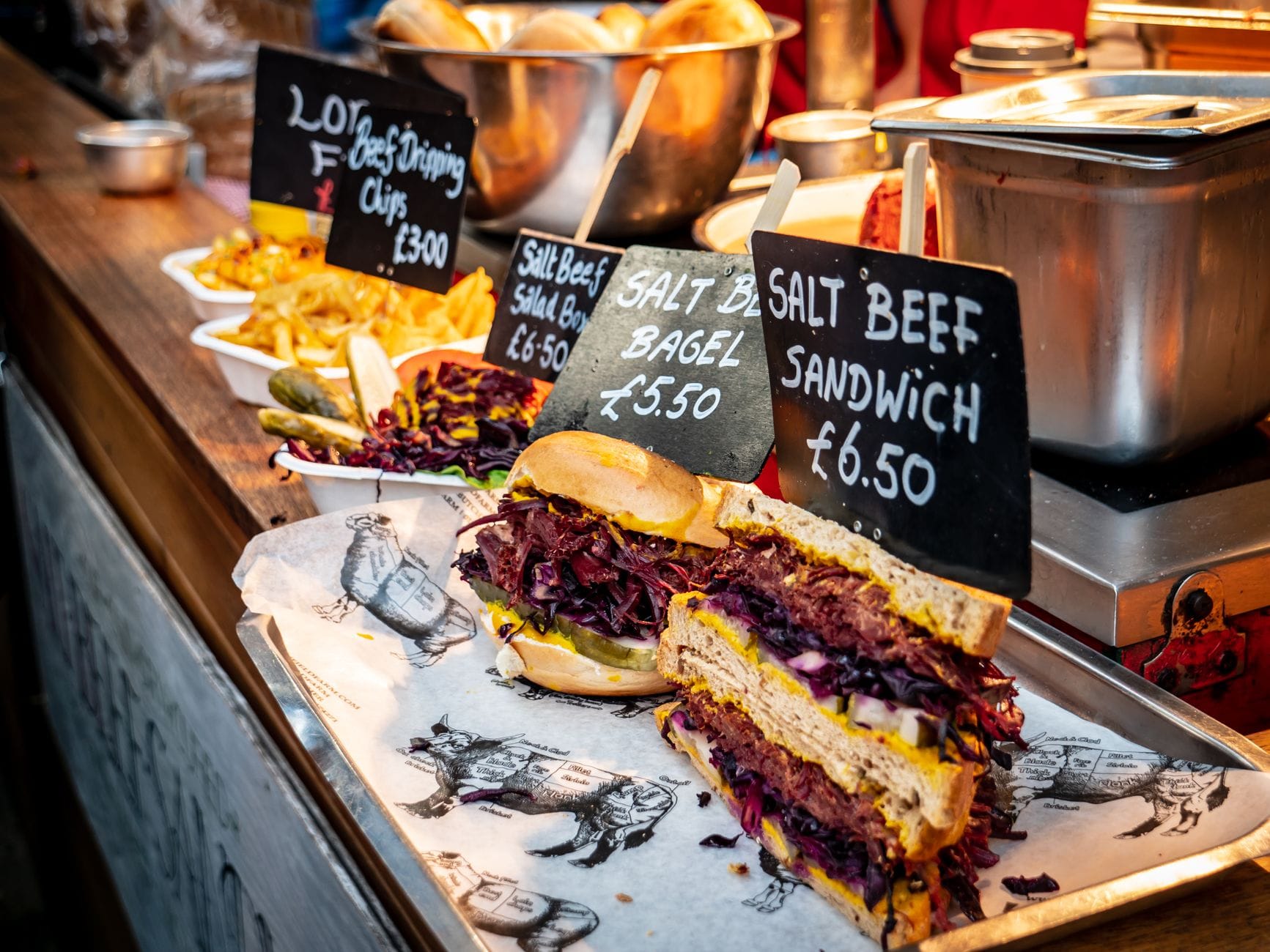 Close-up of a vibrant food market stall with salt beef sandwiches, chips, and bagels, highlighting diverse street food options and handwritten prices.