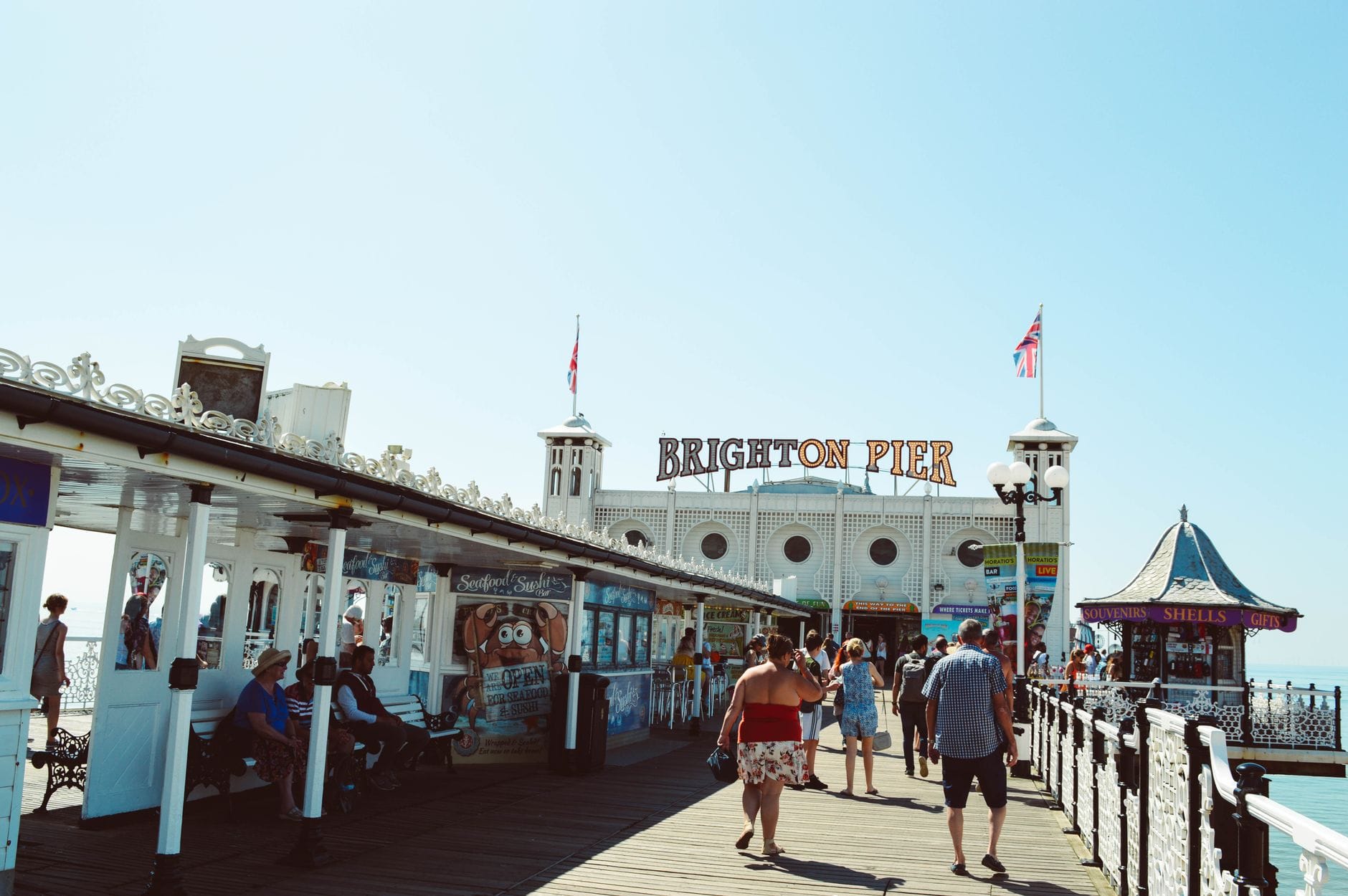 Ingresso al Brighton Pier con persone che camminano, bancarelle di cibo e cartelli, che rappresentano un elemento chiave della cultura balneare e gastronomica di Brighton.
