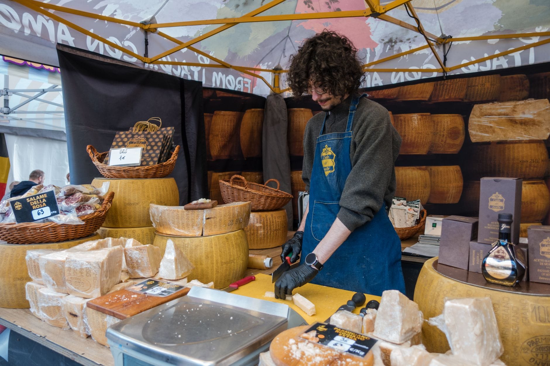 Una bancarella di mercato con un venditore che taglia formaggio su un tavolo affollato, circondato da grandi forme di formaggio e cesti.