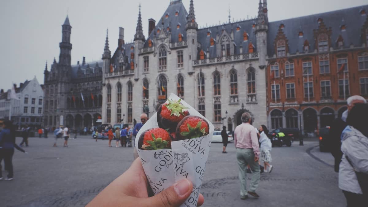 Hand holding a cone of chocolate-covered strawberries in Bruges' main square, with the historic gothic-style city hall and people in the background.