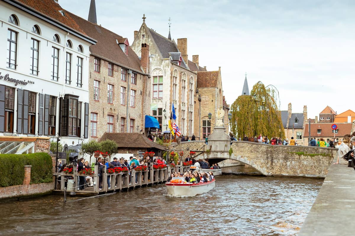 Tourists waiting by a canal for a boat tour in Bruges, with historic buildings and Belgian flags visible along the water.