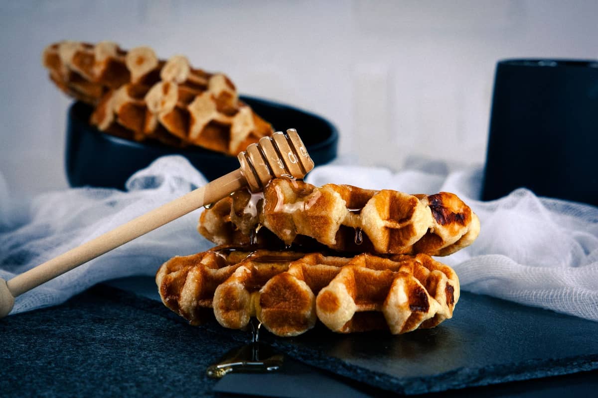 Two Liege style Belgian waffles stacked, drizzled with honey, with another waffle in the background on a dark slate plate.