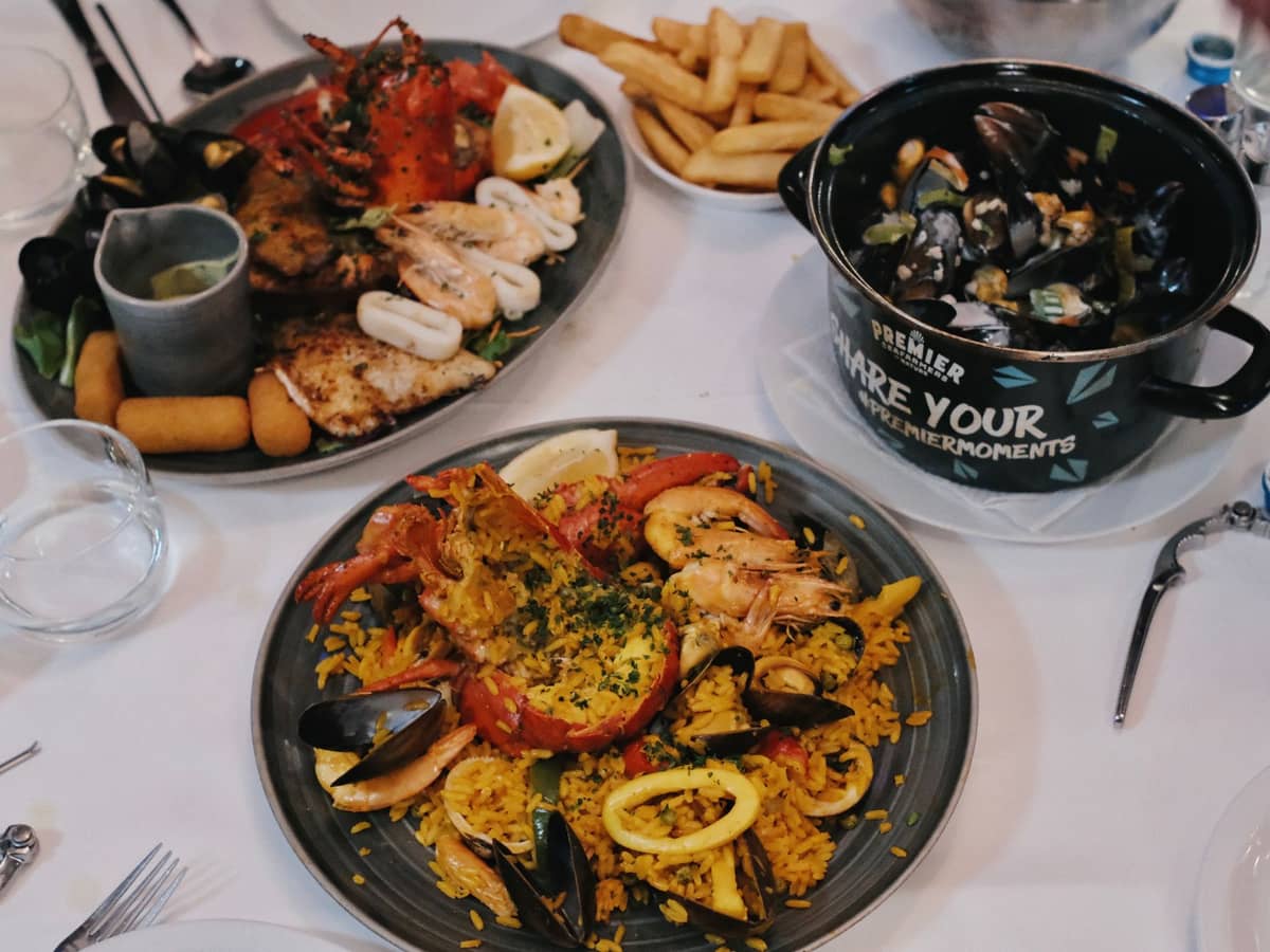 Uma mesa de restaurante com moules-frites (mexilhões e batatas fritas) numa panela, frutos do mar grelhados, croquetes e frites.