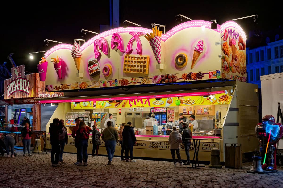 Barraca de comida noturna colorida decorada com waffles, casquinhas de sorvete e outras guloseimas, com pessoas em fila em uma praça de uma cidade belga.