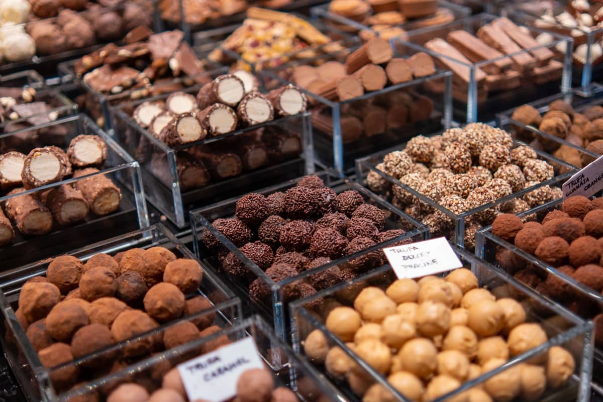 Exposição de bombons de chocolate sortidos e trufas em caixas transparentes numa loja de chocolataria, mostrando variedade e qualidade artesanal.