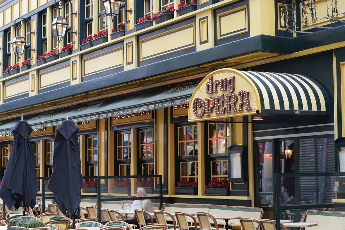 Vista exterior da colorida brasserie 'Drug Opera' em Bruxelas com esplanada e toldo listrado, sugerindo um bairro gastronômico local.