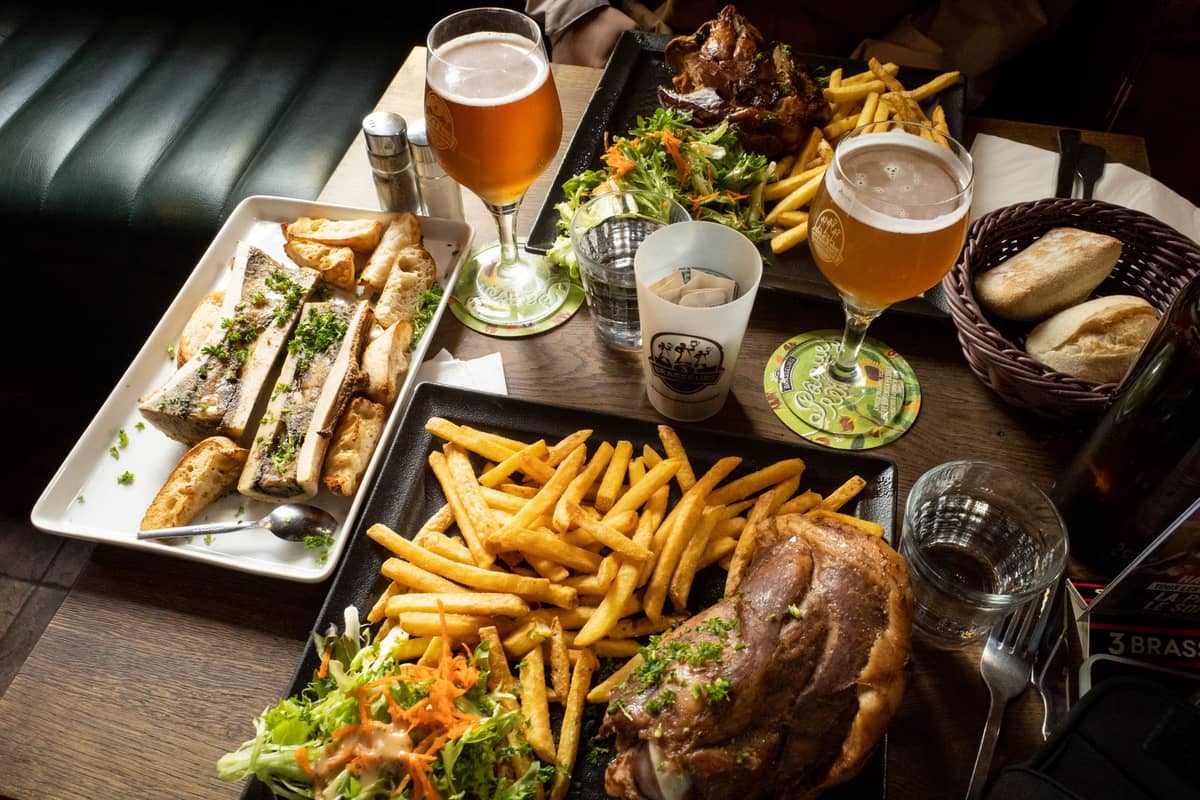 Pratos da culinária belga com batatas fritas, salada, carne e tutano acompanhados de copos de cerveja belga em uma mesa de restaurante.