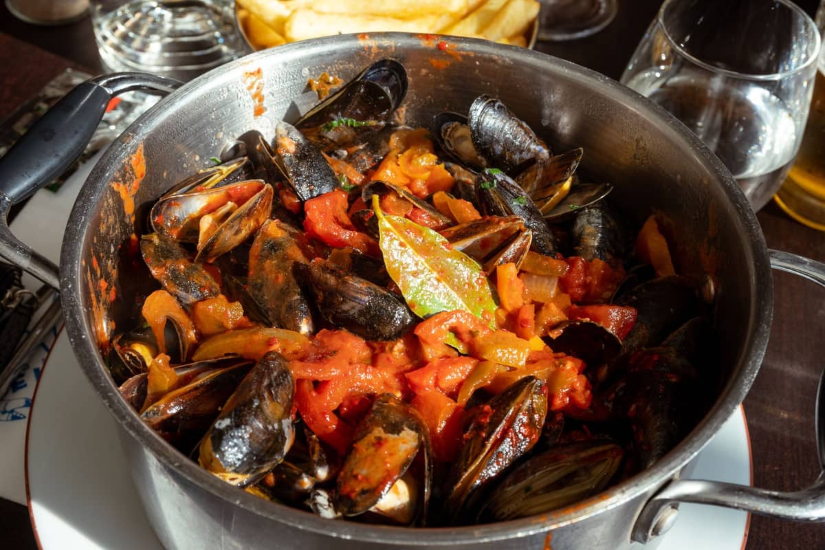 Uma panela de moules-frites (mexilhões com legumes) sobre uma mesa de restaurante à luz do dia, rodeada de copos, batatas fritas e bebidas.