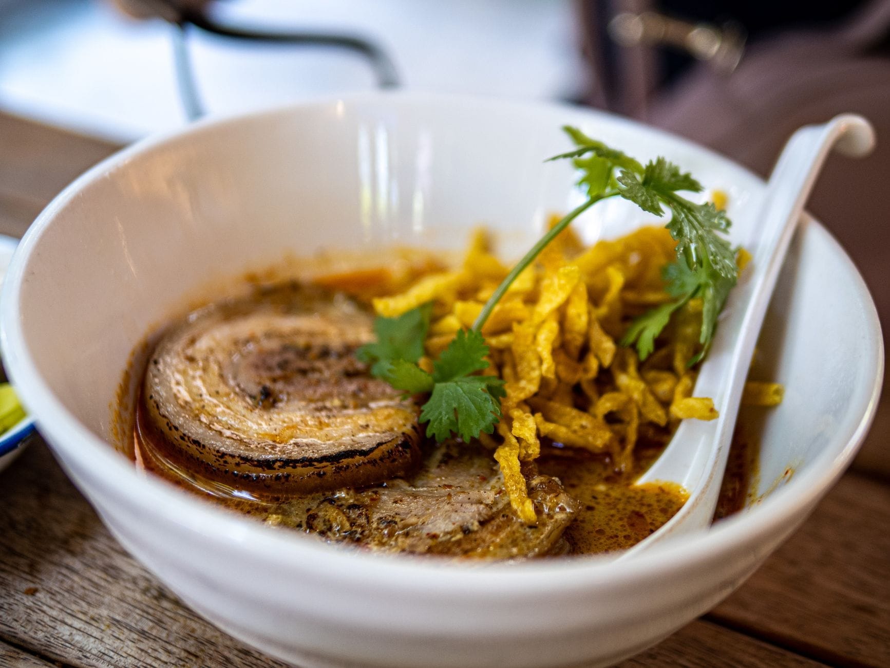 Một tô khao soi với nước dùng cà ri dừa, mì giòn, thịt và rau mùi tươi trên bàn gỗ.