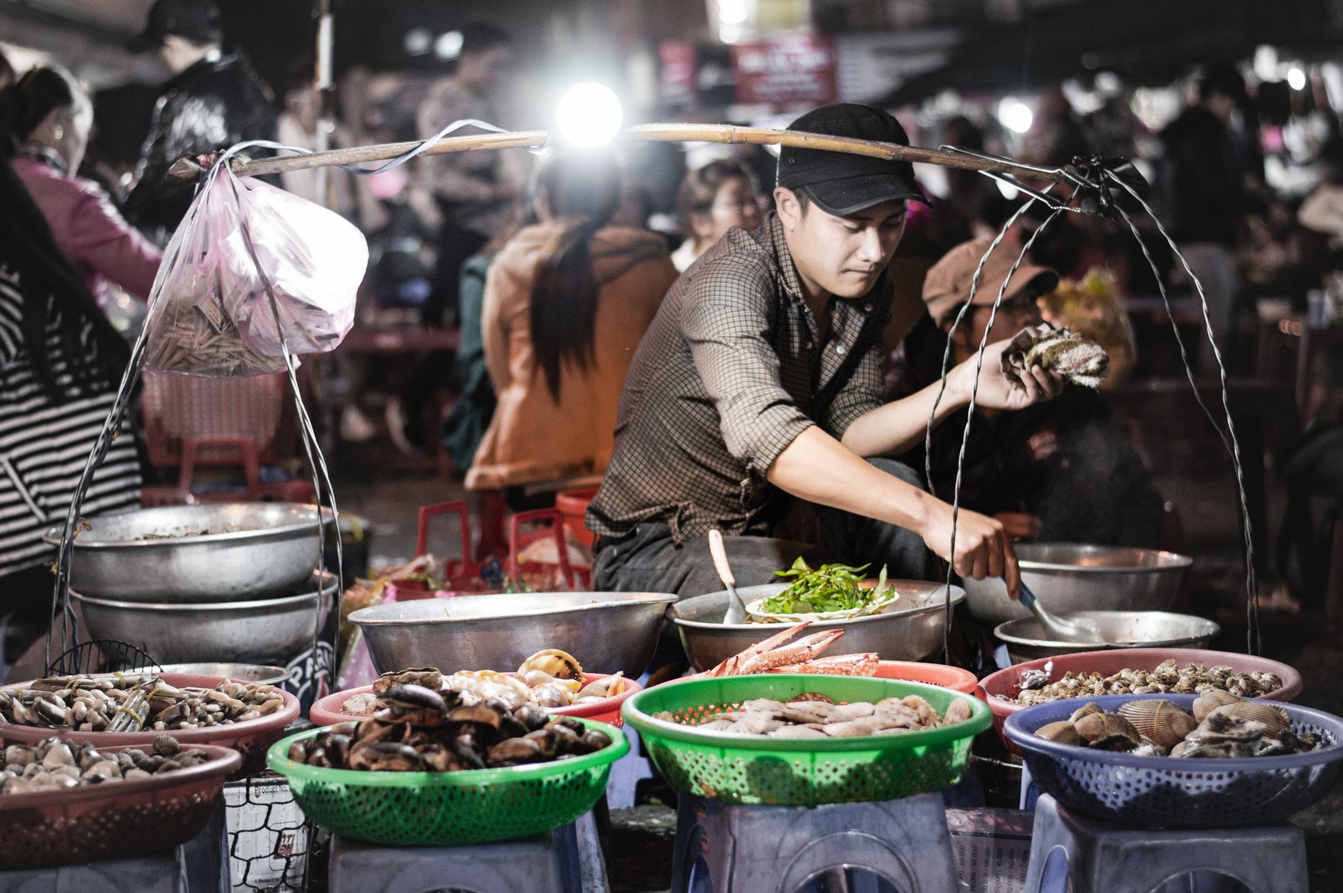 แผงอาหารในตลาดกลางคืนที่เวียดนาม มีพ่อค้าแม่ค้ากำลังเตรียมอาหารทะเลอยู่หลังตะกร้าหอยและสัตว์น้ำมีเปลือก บรรยากาศร้านอาหารริมถนน