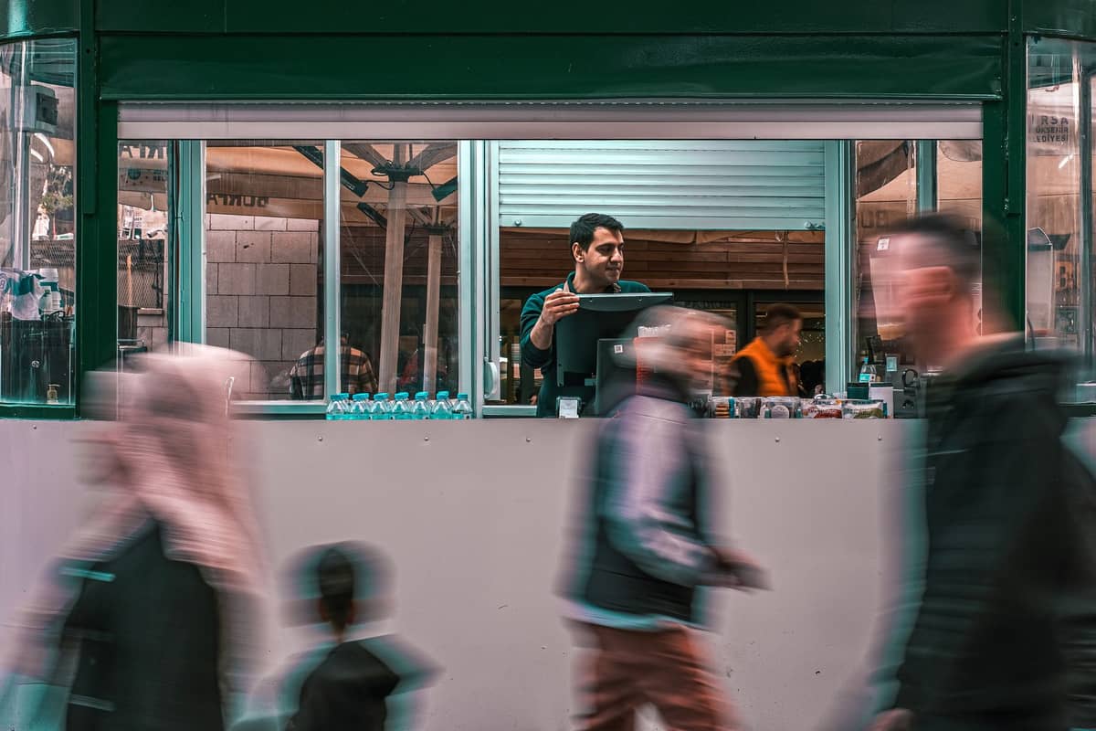 Straßenverkäufer in einem grünen Kiosk, lächelnd inmitten verschwommener Passanten, unter hellem Licht, das alltägliches entspanntes Essen in einer nahöstlichen Stadt evoziert.