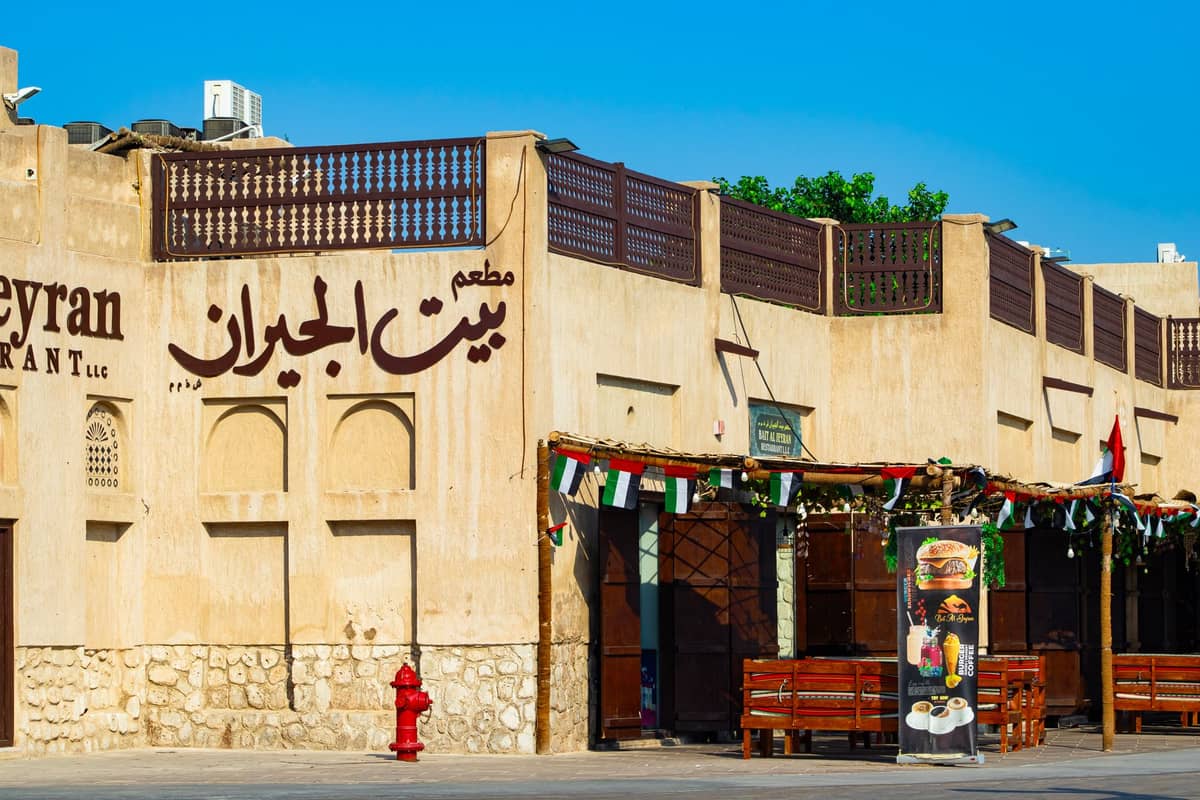 Außenansicht eines traditionell wirkenden emiratischen Restaurants in Dubai, mit arabischer Beschilderung, Außensitzplätzen und dekorativen Flaggen unter einem klaren blauen Himmel.
