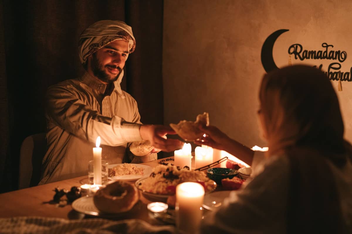 Ein Mann und eine Frau teilen Essen an einem kerzenbeschienenen Tisch, der mit traditionellen Gerichten gedeckt ist, mit Ramadan-Grüßen an der Wand hinter ihnen.