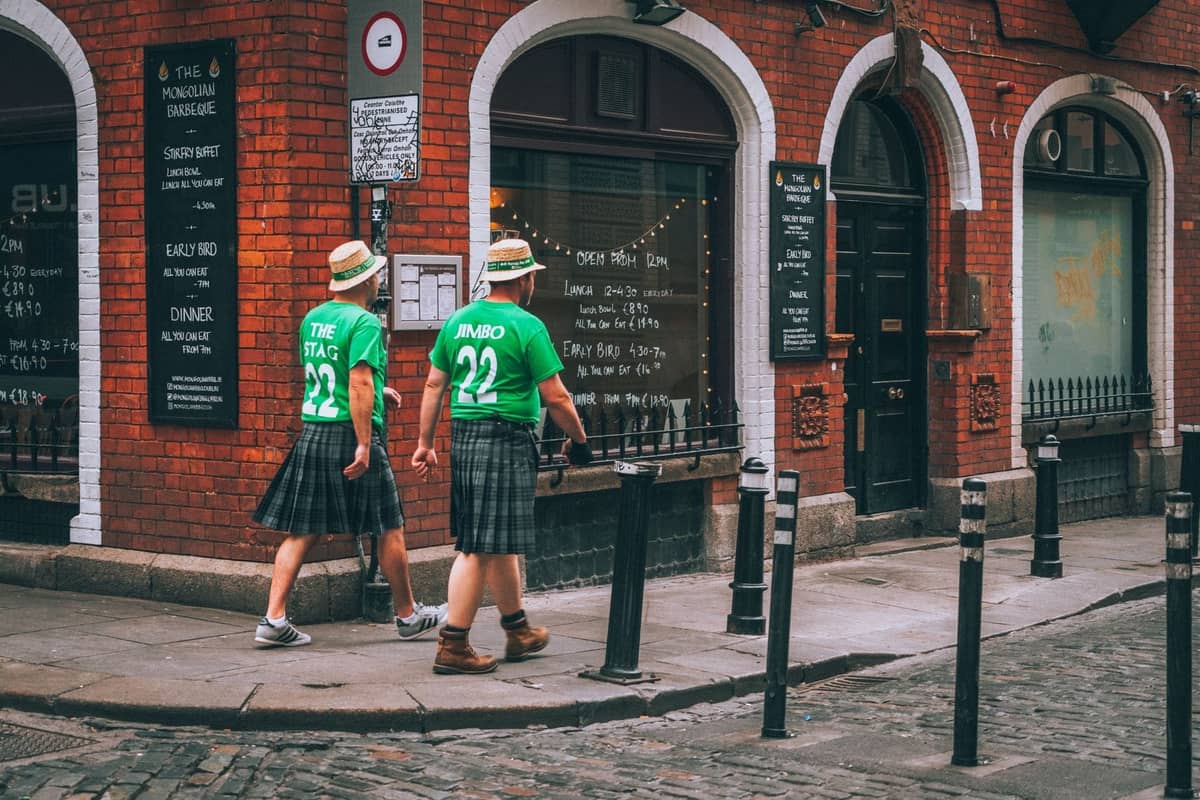 Due persone in kilt e magliette verdi camminano davanti a un pub di Dublino con menu scritti a mano sulle finestre.
