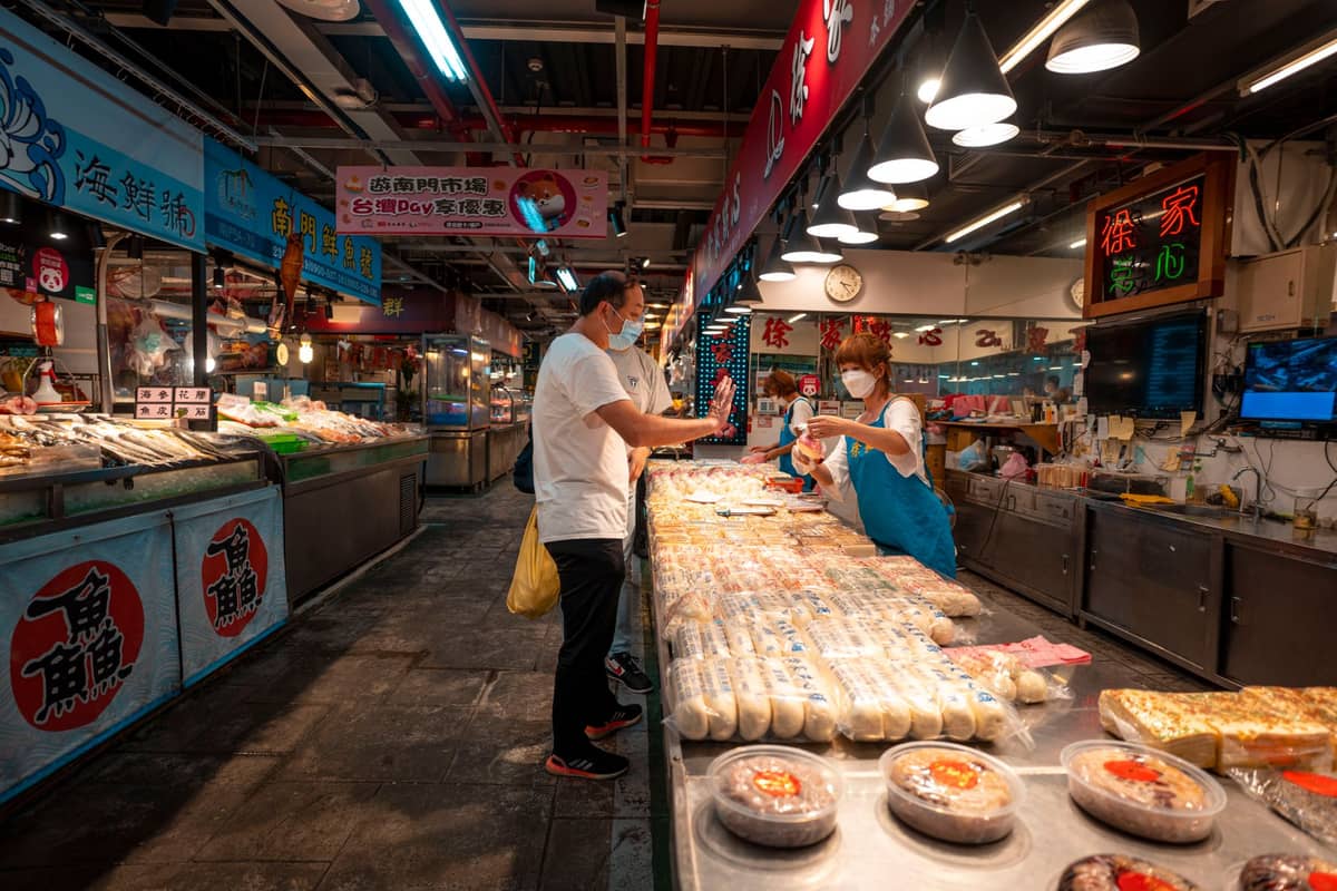 Indoor market in Guangzhou with vendor handing food to a customer, showing a variety of fresh Cantonese ingredients under market signs.