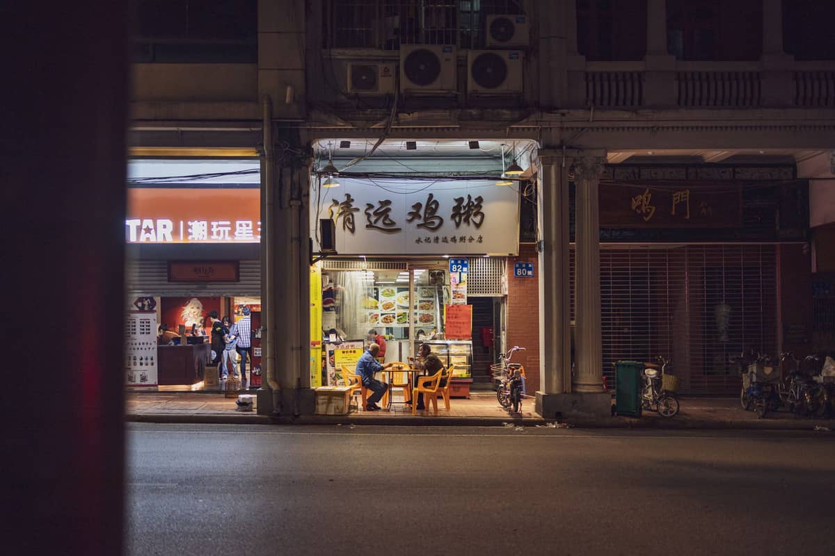 Nighttime street view of a small, traditional Cantonese restaurant with a lit sign in Chinese and people eating inside.