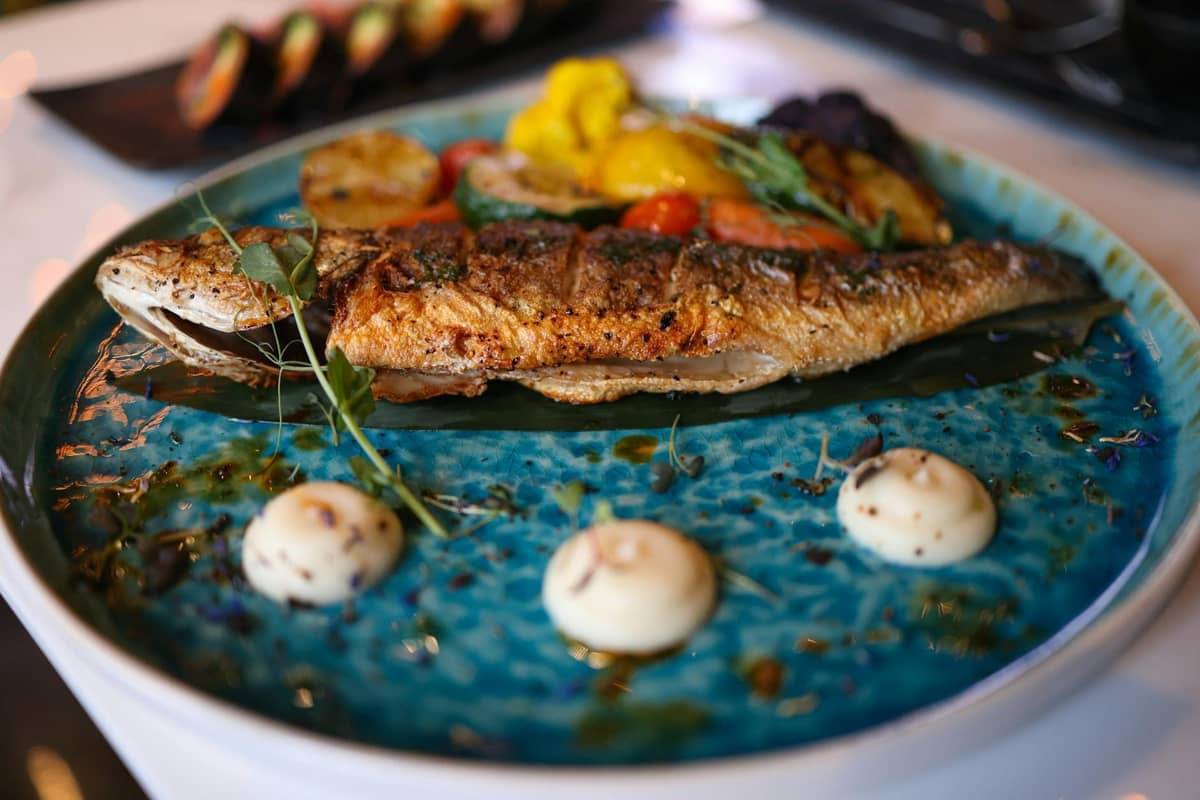 A whole grilled fish served on a blue ceramic plate with a garnish of vegetables, beautifully presented in a restaurant setting.
