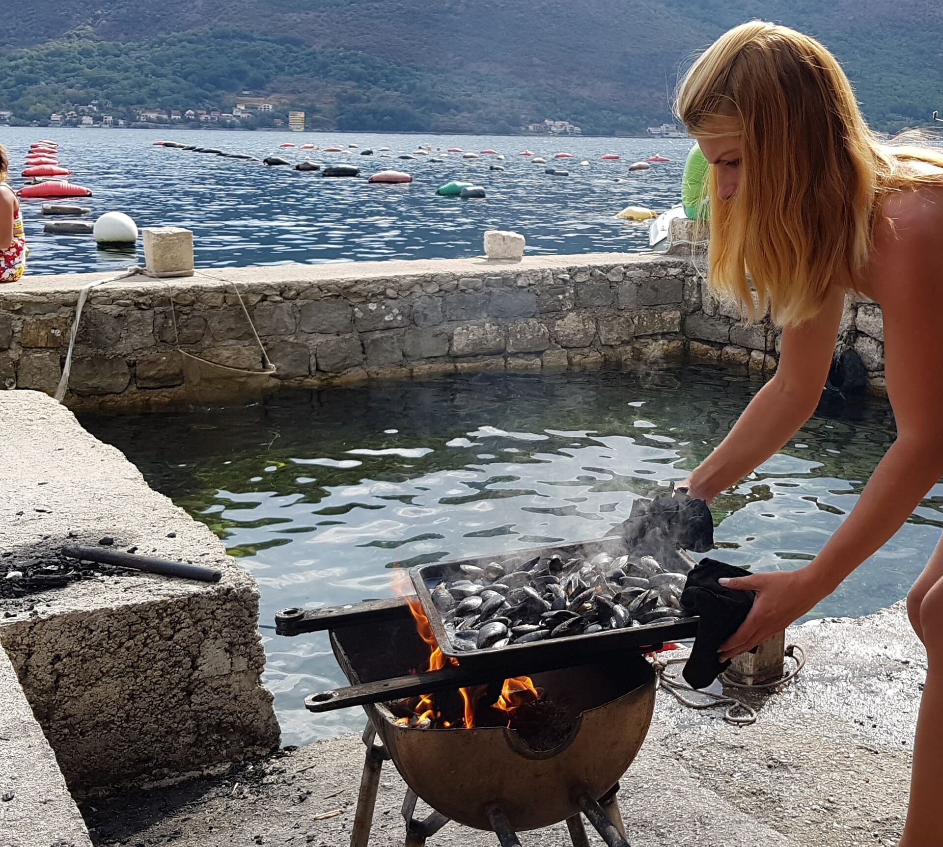 Uma mulher cozinha mexilhões frescos sobre chama aberta à beira-mar em Kotor, com a Baía de Boka e as montanhas ao fundo.