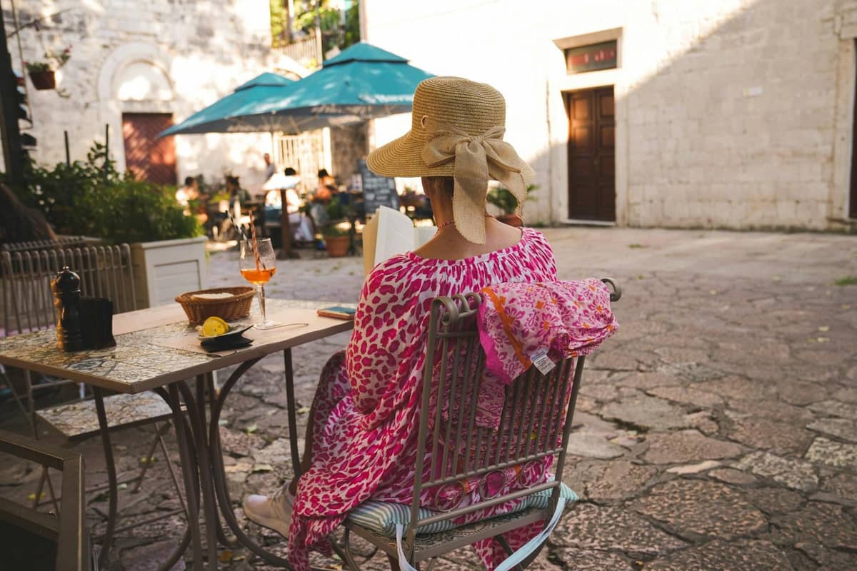 Woman in a straw hat sitting at an outdoor cafe table in Kotor with an Aperol spritz visible, stone square background.
