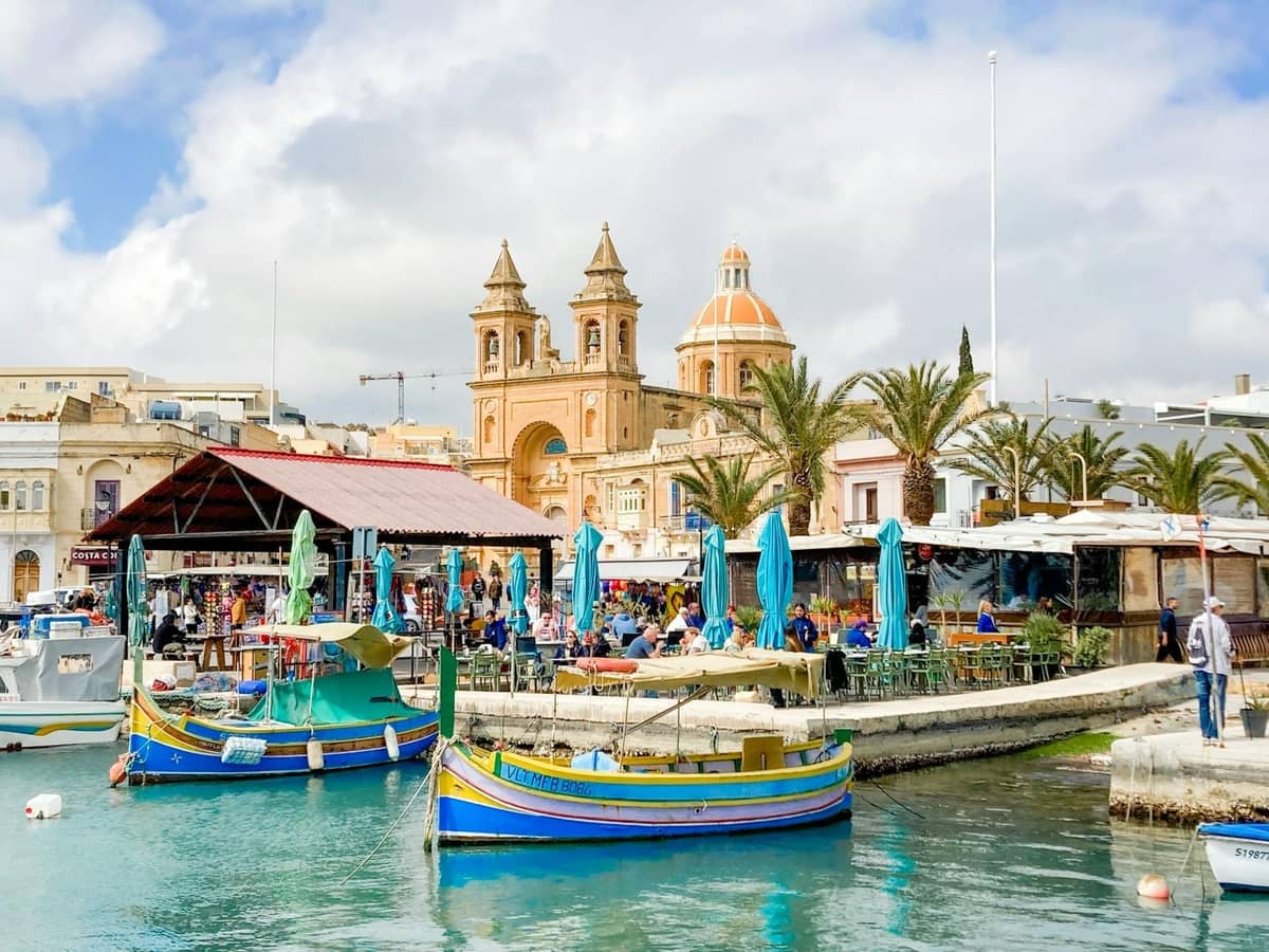 Vivace lungomare maltese con colorati pescherecci, ristoranti all'aperto e una chiesa prominente, che cattura un'autentica scena di ristorazione e mercato.