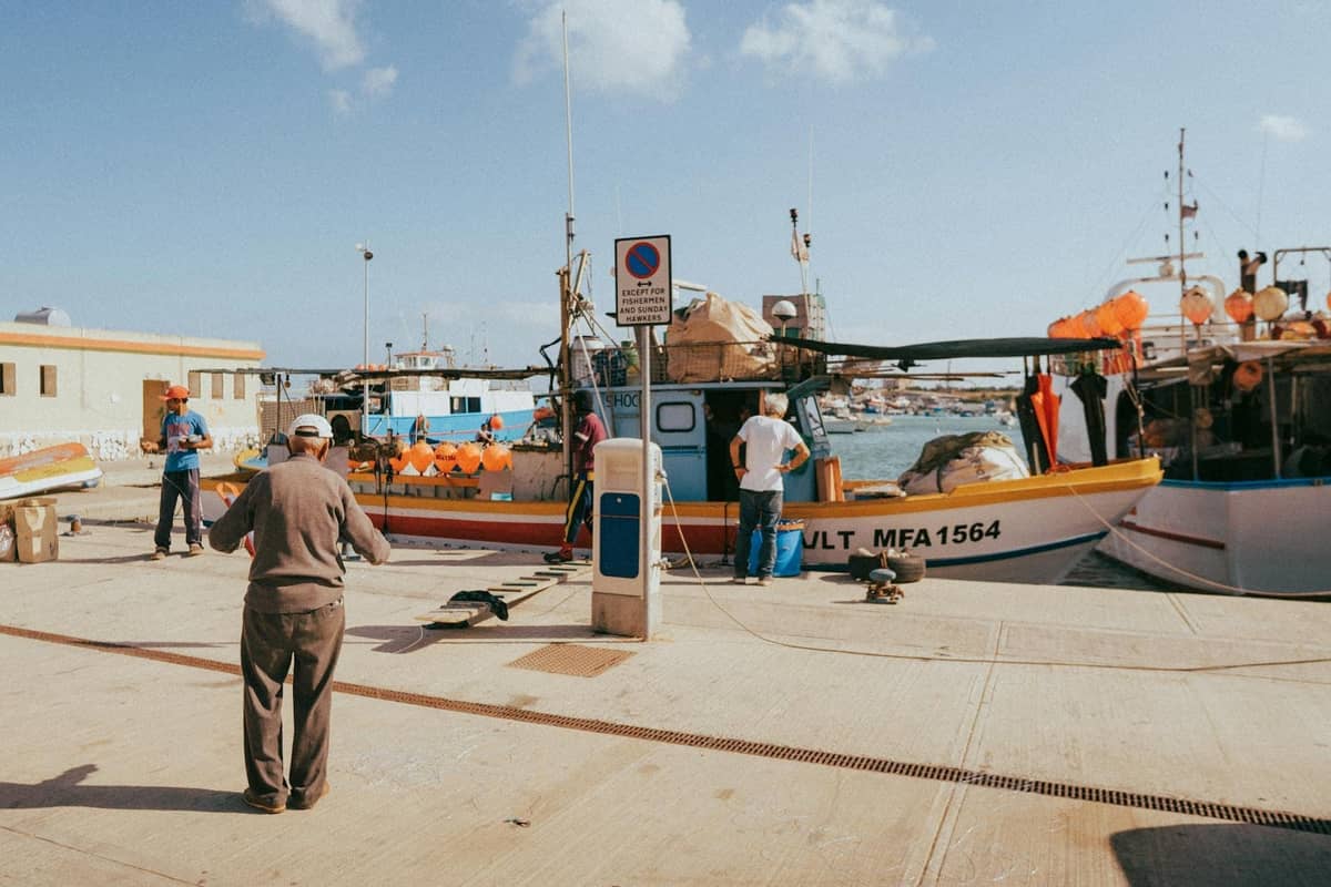 Pescatori in un porto maltese che preparano le barche, con attrezzi da pesca e banchine visibili sotto cieli soleggiati.