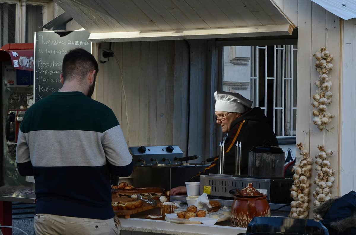 Un uomo che acquista pasticcini da un piccolo chiosco di street food dove una persona con cappello da chef sta servendo. I pasticcini sono esposti in primo piano.