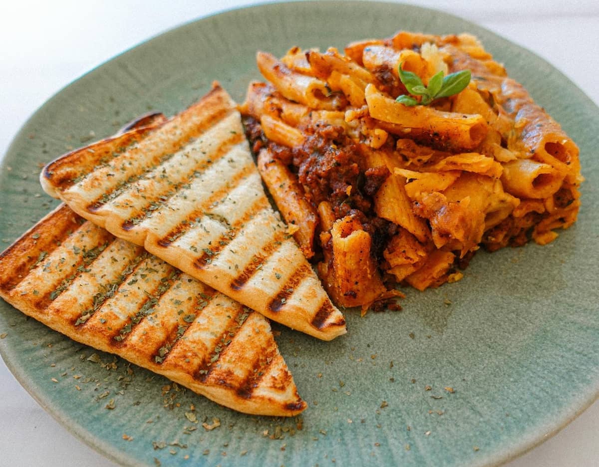 Piatto di rigatoni al forno con sugo di carne, servito con due pezzi di pane grigliato.
