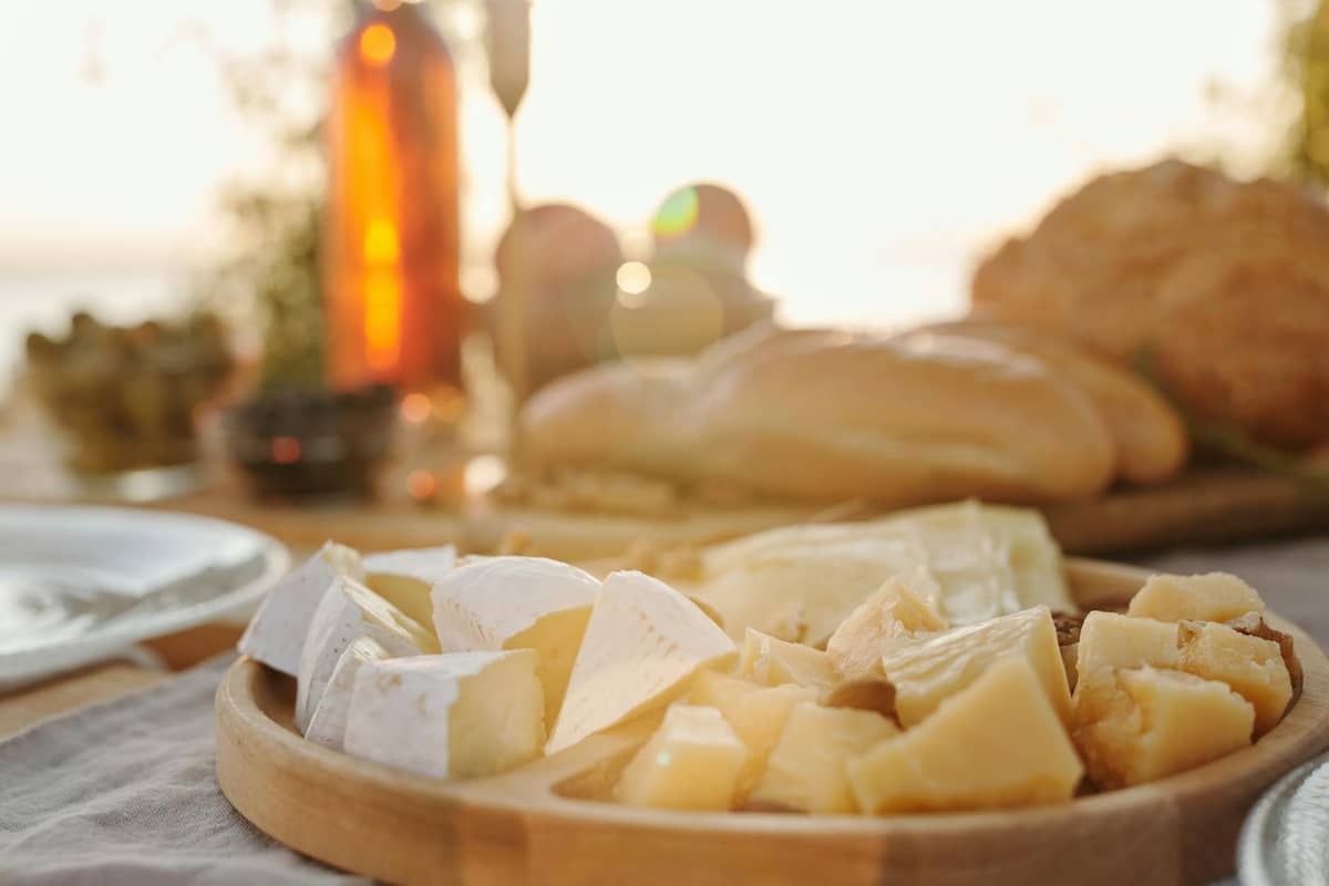 Vassoio di legno con formaggi assortiti, pane, olive e bottiglie di vino su un tavolo all'aperto alla luce del sole, che evoca un'atmosfera mediterranea