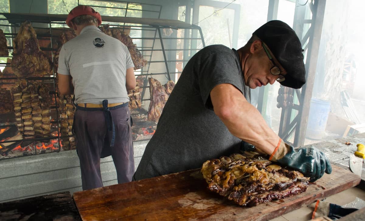 ชายสองคนกำลังเตรียม asado แบบอุรุกวัย คนหนึ่งแล่เนื้อย่างบนโต๊ะ อีกคนดูแลซี่โครงและชิ้นเนื้อที่กำลังย่างบนถ่านไม้ใน parrilla แบบดั้งเดิม