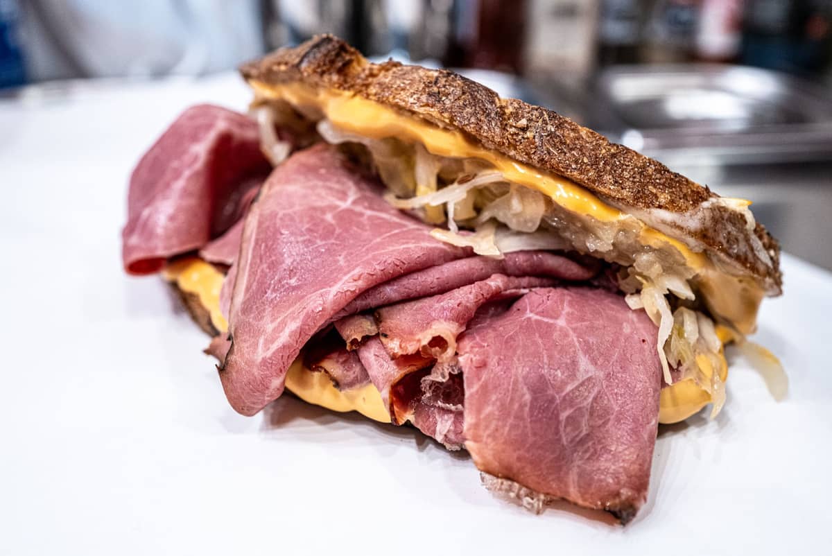 Nahaufnahme eines herzhaften Sandwichs mit dicken Scheiben Räucherfleisch, geschmolzenem Käse und Sauerkraut auf Roggenbrot, angerichtet auf einem weißen Teller.