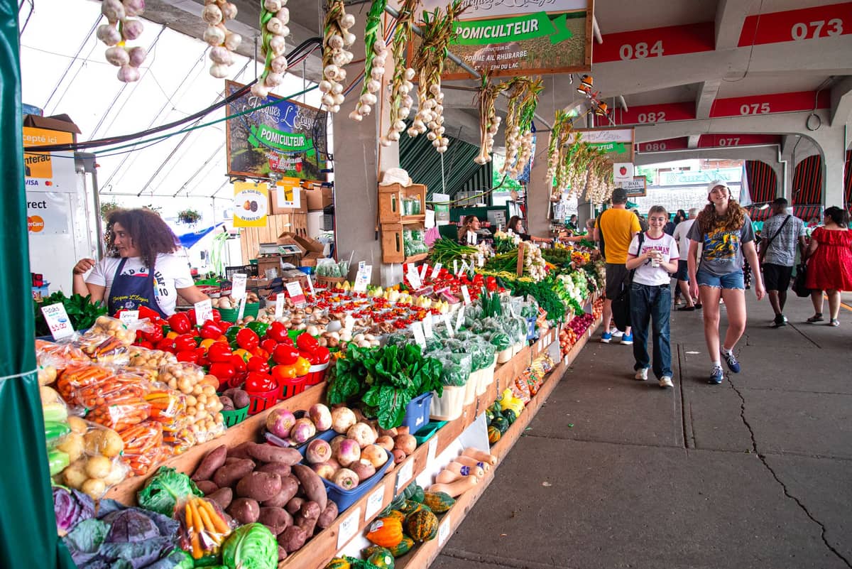 Ein lebhafter Marktstand auf dem Marché Jean-Talon in Montreal mit frischem Gemüse, Obst und Einkäufern, die unter einer überdachten Struktur vorbeigehen.