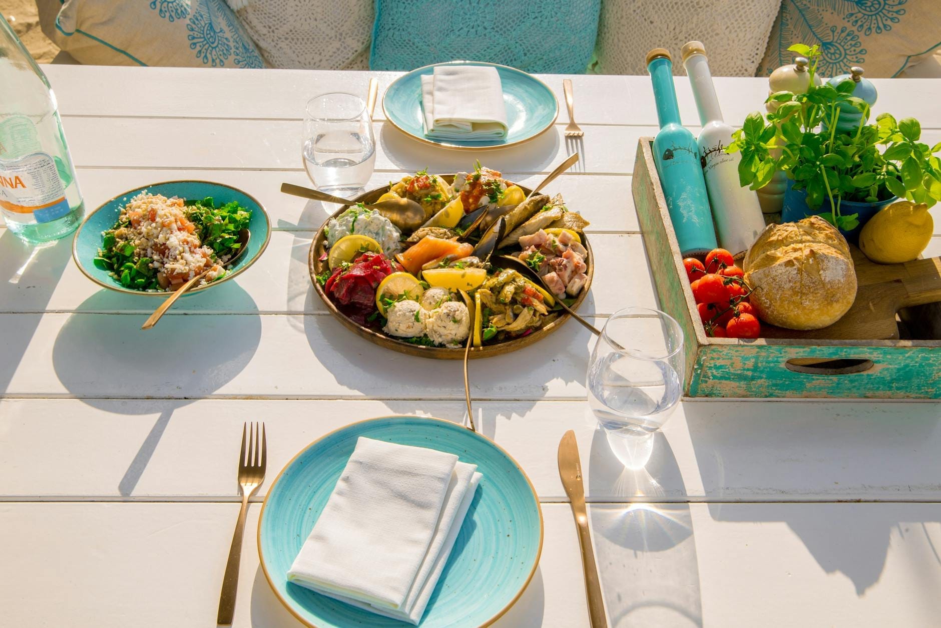 Colorida bandeja griega de mezze, ensalada, pan, hierbas y bebidas sobre una mesa blanca iluminada por el sol en Mykonos, con platos turquesa y cómodos cojines.