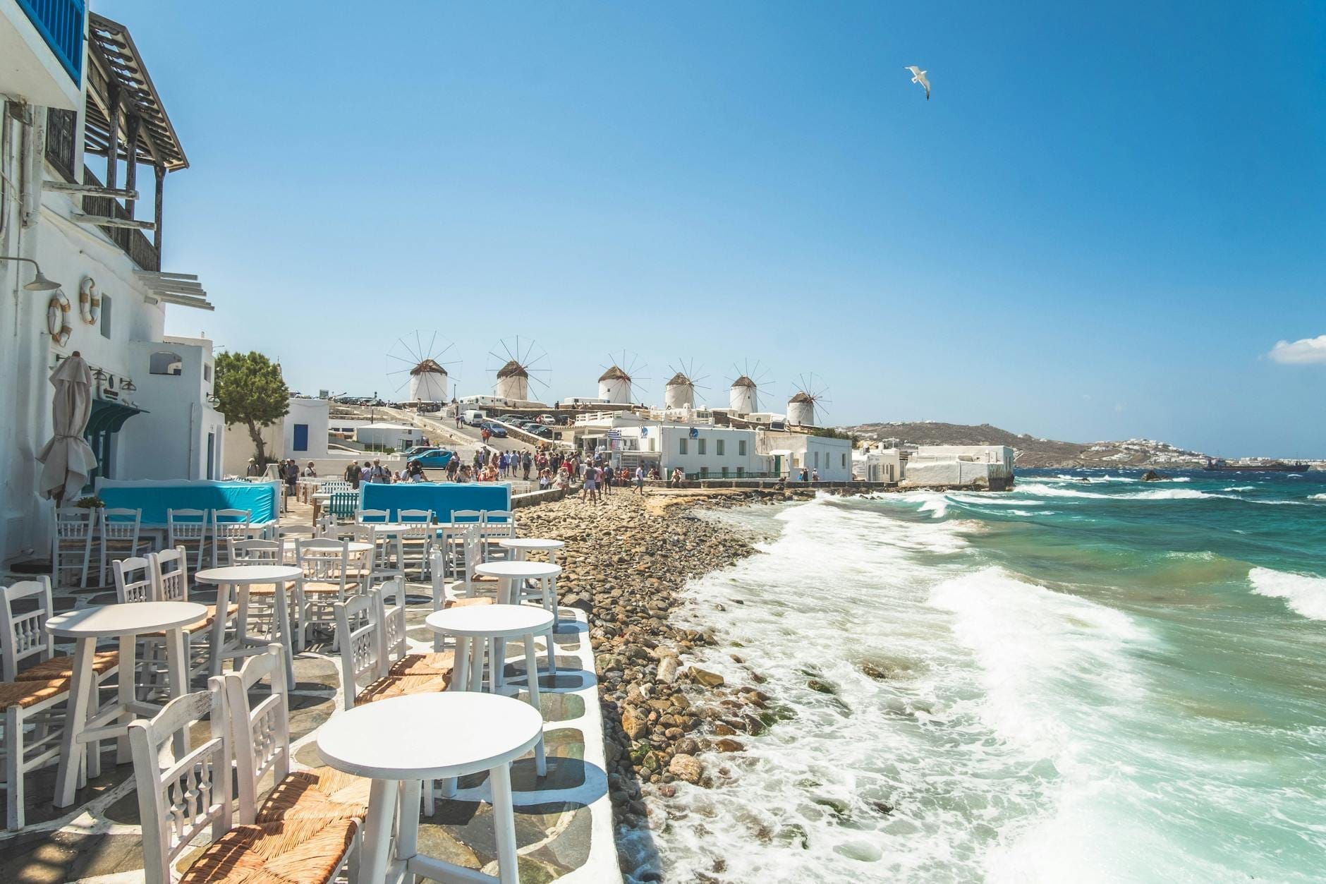 Mesas de restaurante frente al mar en Mykonos con vistas a los famosos molinos de viento y aguas turquesas, ilustrando la alta demanda de reservas durante la temporada turística alta.