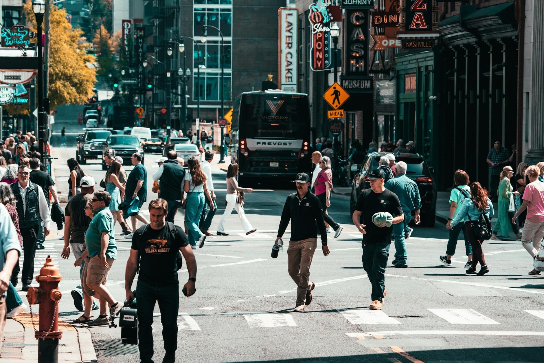 Concurrida escena en un paso de peatones en el centro de Nashville con personas caminando, letreros de neón y clásicos locales de música al fondo.