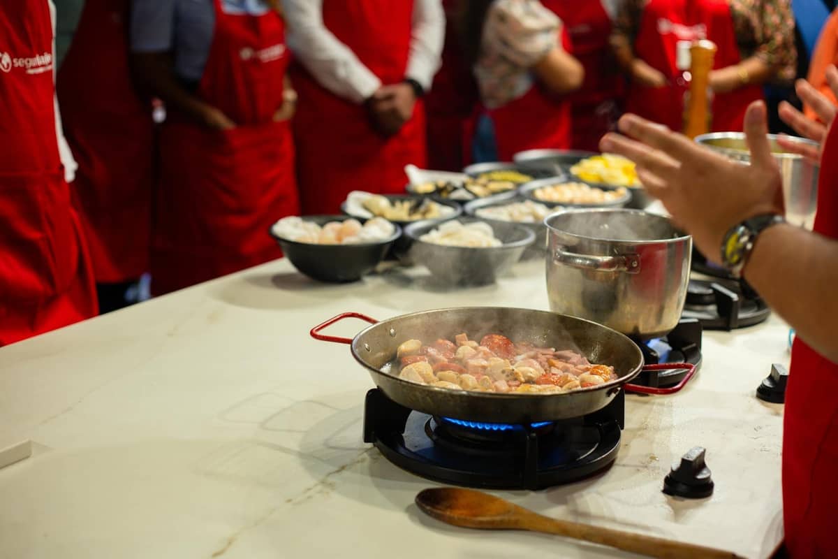 Cooking class scene with multiple people in red aprons watching as someone prepares traditional food in a pan, illustrating shared culinary traditions.