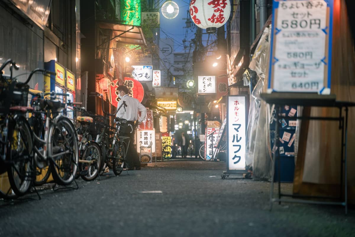 Neon-lit Osaka food alley at night with restaurant signs, bicycles, and people walking, evoking a typical street food tour scene.